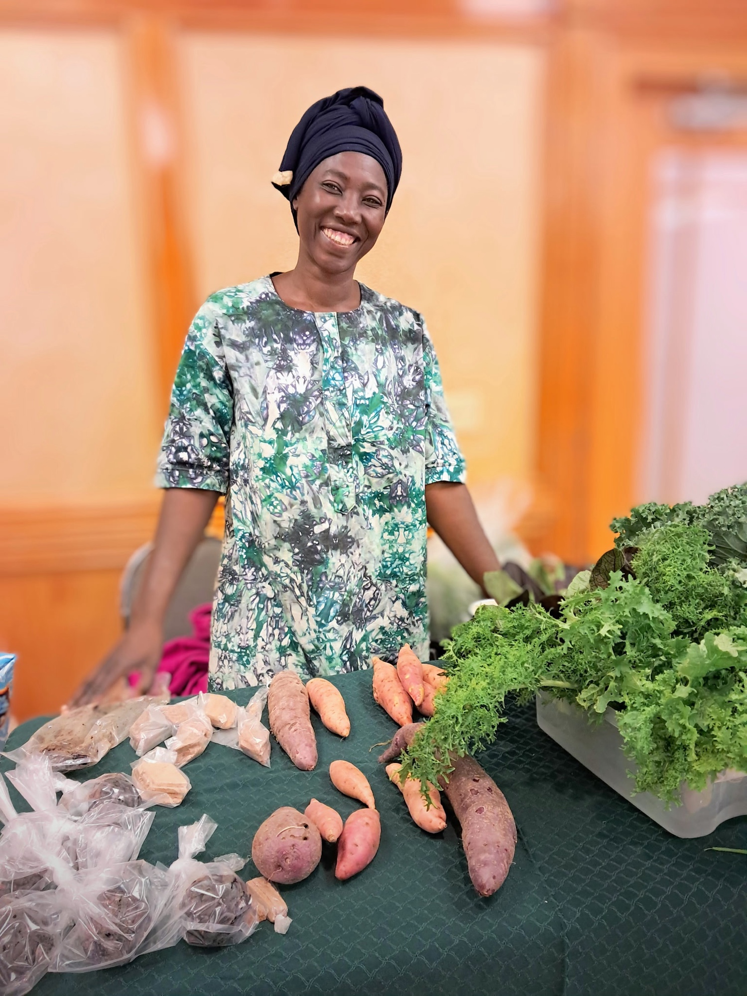 Francisca Hien smiling at her market table with sweet potatoes, kale, mustard greens, and herbs